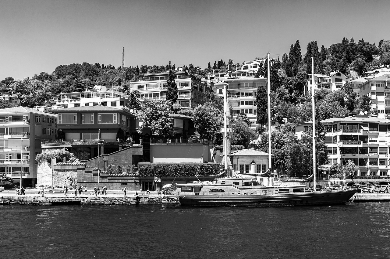 モノクロームで捉えられたボスポラス海峡沿いの風景。海沿いに係留された木造の大型ヨットと、緑豊かな斜面に建ち並ぶモダンな邸宅や洋館群が見える。